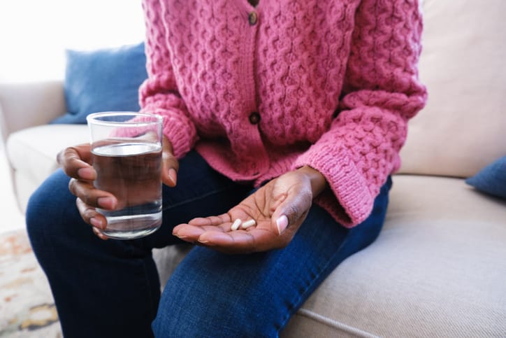 Photo shows a woman holding pills and a glass of water on the couch/Getty Images