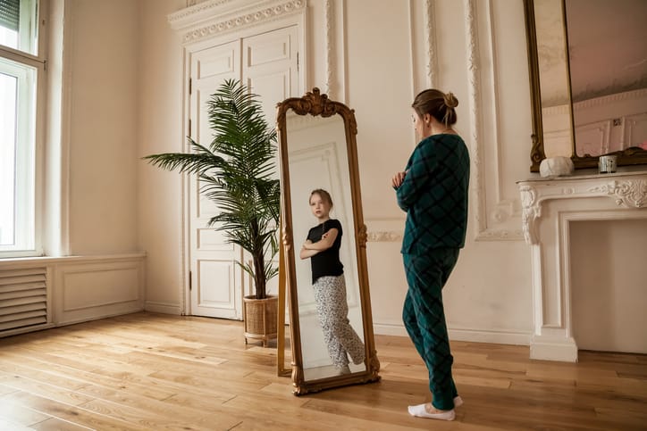 Photo shows an adult woman seeing her childhood self in a mirror/Getty Images